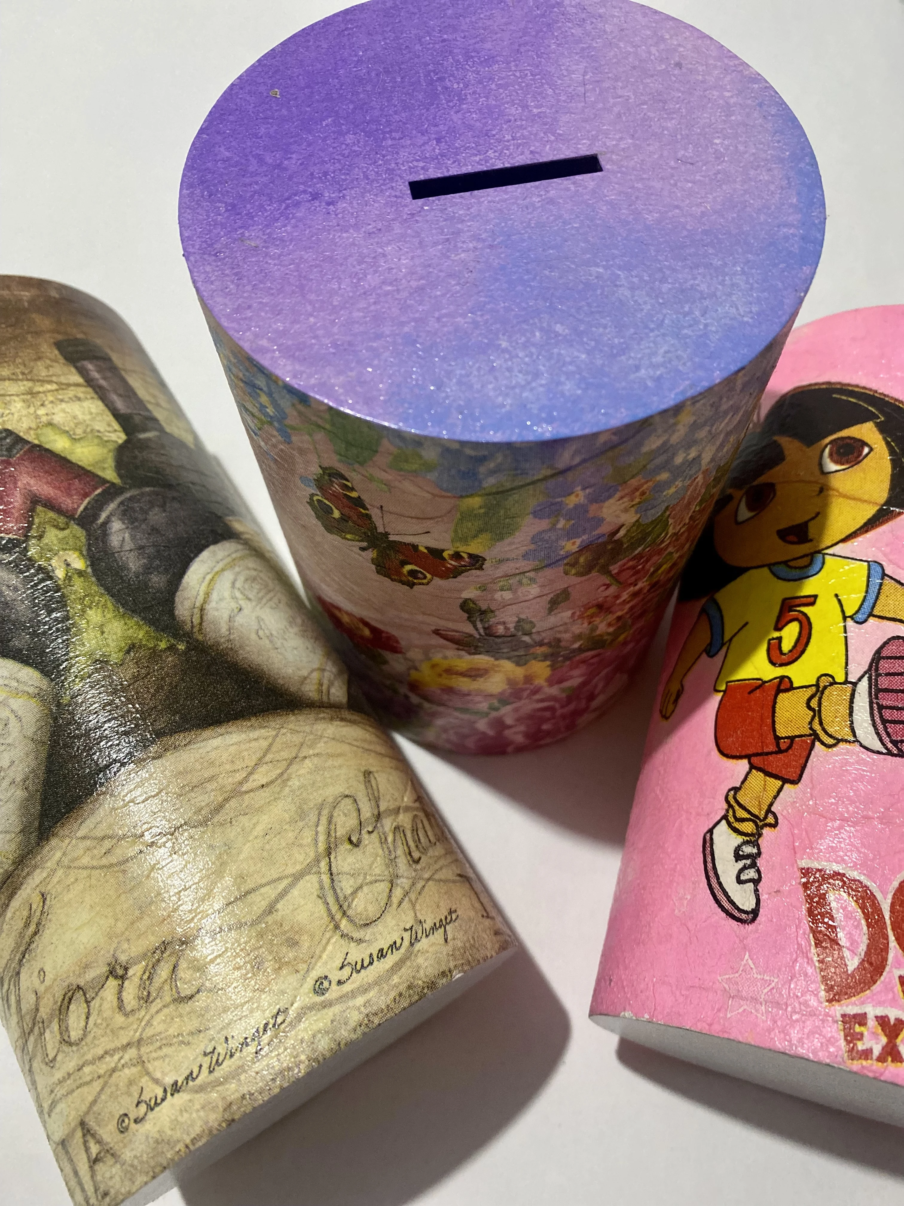 Tres alcancías cilíndricas: una con diseño de vino y firma Susan Winget, una floral con tapa morada, y una rosa de personaje infantil.