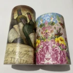 Dos alcancías cilíndricas decoradas: una con botellas de vino estilo rústico y otra con flores vibrantes y mariposa.