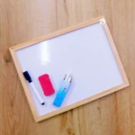 Tablero didáctico blanco con marco de madera, marcador azul, borrador rojo y estuche azul con gis.