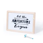 Tablero didáctico blanco con marco de madera clara y frase "Let the adventure begin" escrita en negro.
