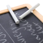 Pizarra didáctica con dos tizas blancas apoyadas, letras escritas con tiza sobre superficie negra.