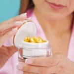 Manos sostienen frasco de Lyralá con perlas doradas de sérum, fondo azul y ropa rosa.