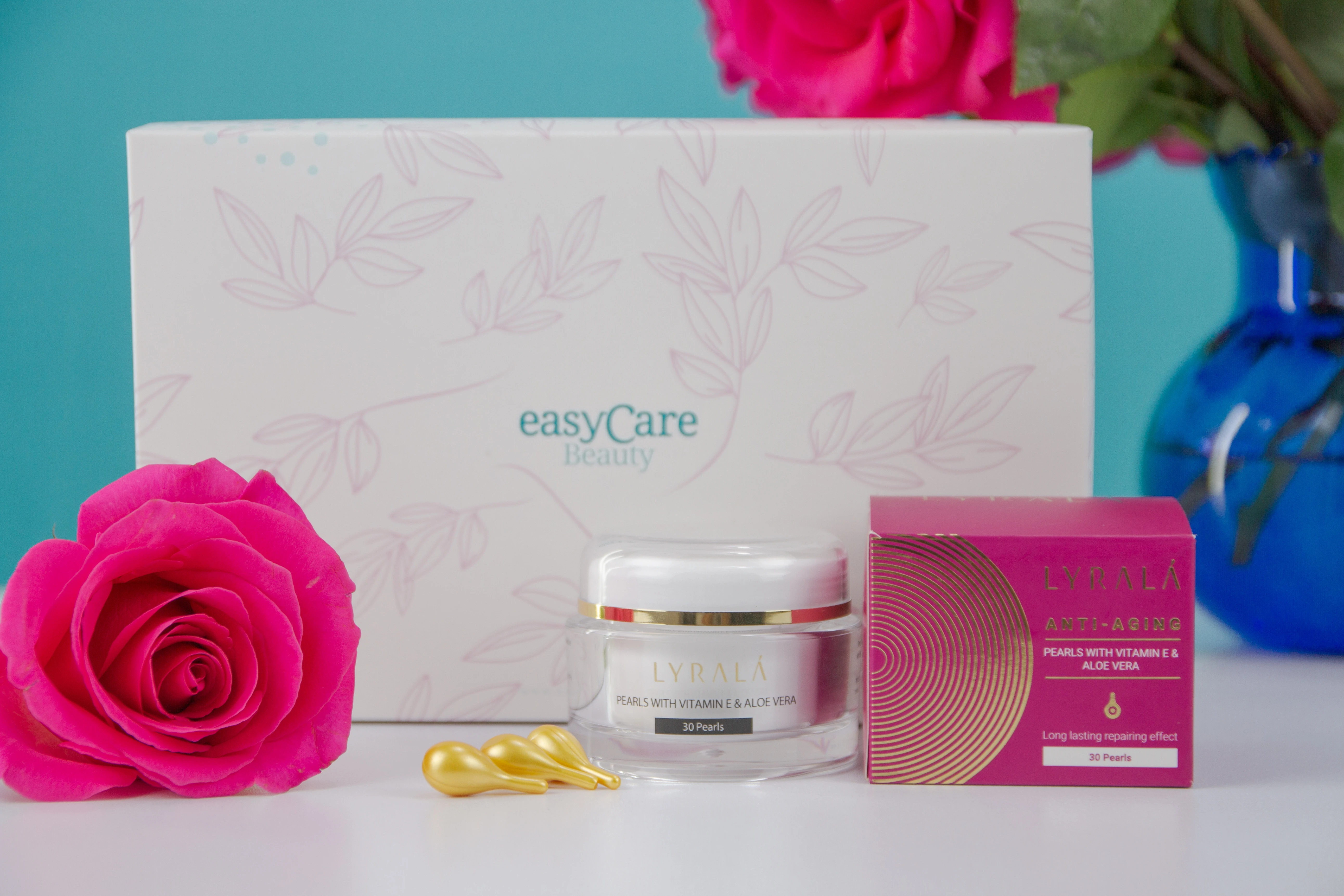 Perlas antiedad Lyralá con Vitamina E, envase blanco con hojas rosas, junto a una rosa fucsia y tres cápsulas doradas.