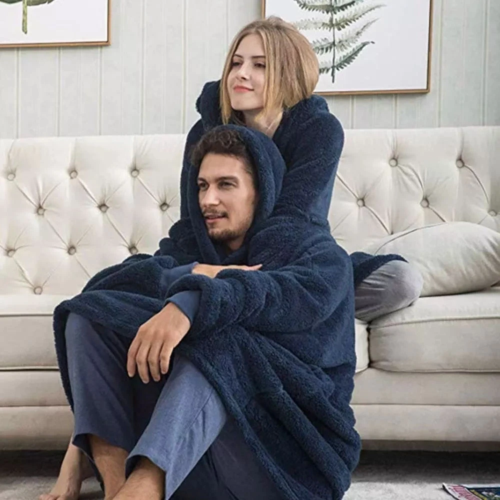 Pareja usando manta térmica azul marino extragrande con capucha, acurrucados en sofá.