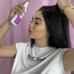 Mujer aplica tónico capilar en spray en su cabello oscuro, de perfil, frente a fondo rosa.