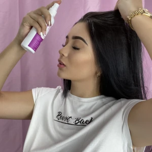 Mujer aplica tónico capilar en spray en su cabello oscuro, de perfil, frente a fondo rosa.