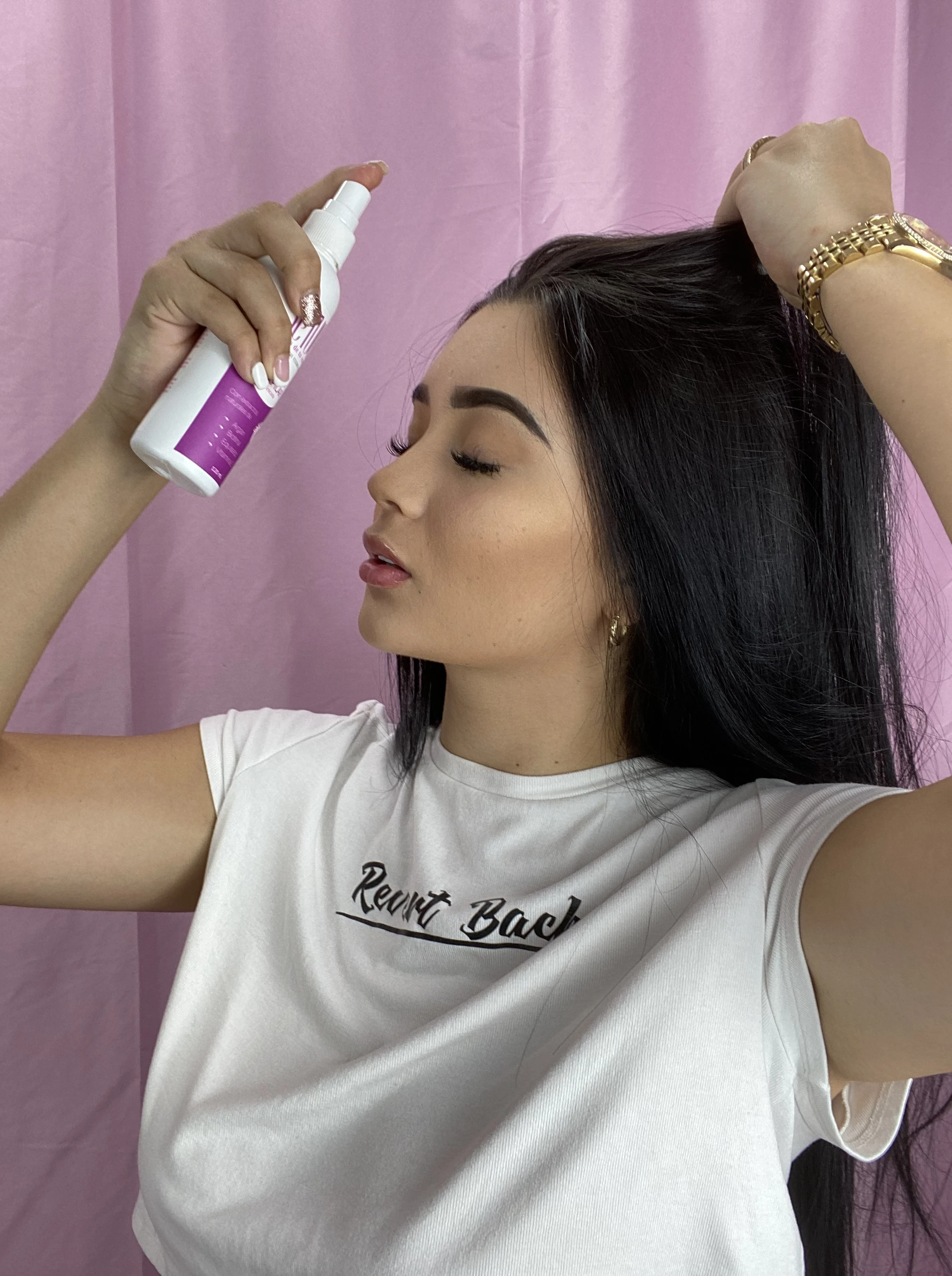 Mujer aplica tónico capilar en spray en su cabello oscuro, de perfil, frente a fondo rosa.