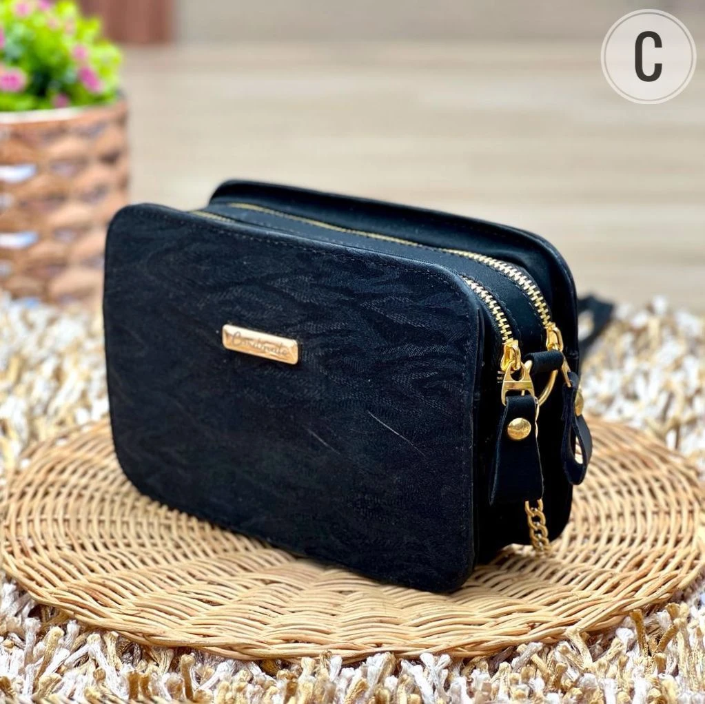 Bolso de mano dama negro, textura sutil, con herrajes y cremalleras doradas, puesto sobre estera de mimbre.