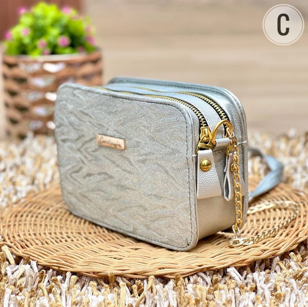 Bolso dama cuadrado plateado con textura animal print sutil, cremallera dorada y cadena pequeña, sobre base de mimbre.