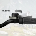 Timbre negro para bicicleta con rotación 360°, montado en manubrio junto a freno, fondo desenfocado de ciclista en montaña.