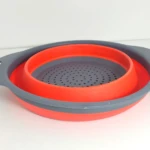 Colador redondo plegable de silicona naranja y gris, con orificios finos y asas.