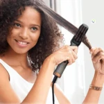 Mujer sonriente usando cepillo alisador Pro950 gris con detalles cobre; sus dientes cerámicos alisan una sección de cabello rizado oscuro desde la raíz.