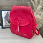 Morral Dama Agatha fucsia con borla dorada, cremalleras y logo, presentado con revista Vogue.