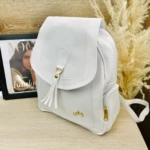 Bolso morral blanco para dama Agatha con tapa, borlas y zíper dorado, sobre mesa de madera.