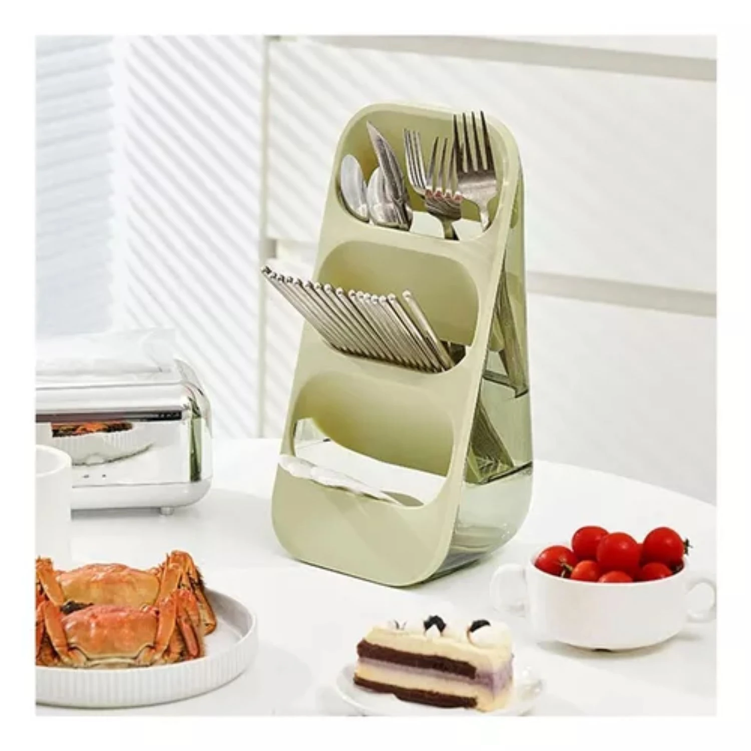Organizador de cubiertos de tres niveles en color verde pastel y base transparente, posicionado sobre una mesa blanca junto a vajilla con comida.