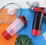Dos termos shaker de proteína, uno transparente y uno negro con bases y tapas rojas, junto a bádminton, béisbol y pesa cromada.