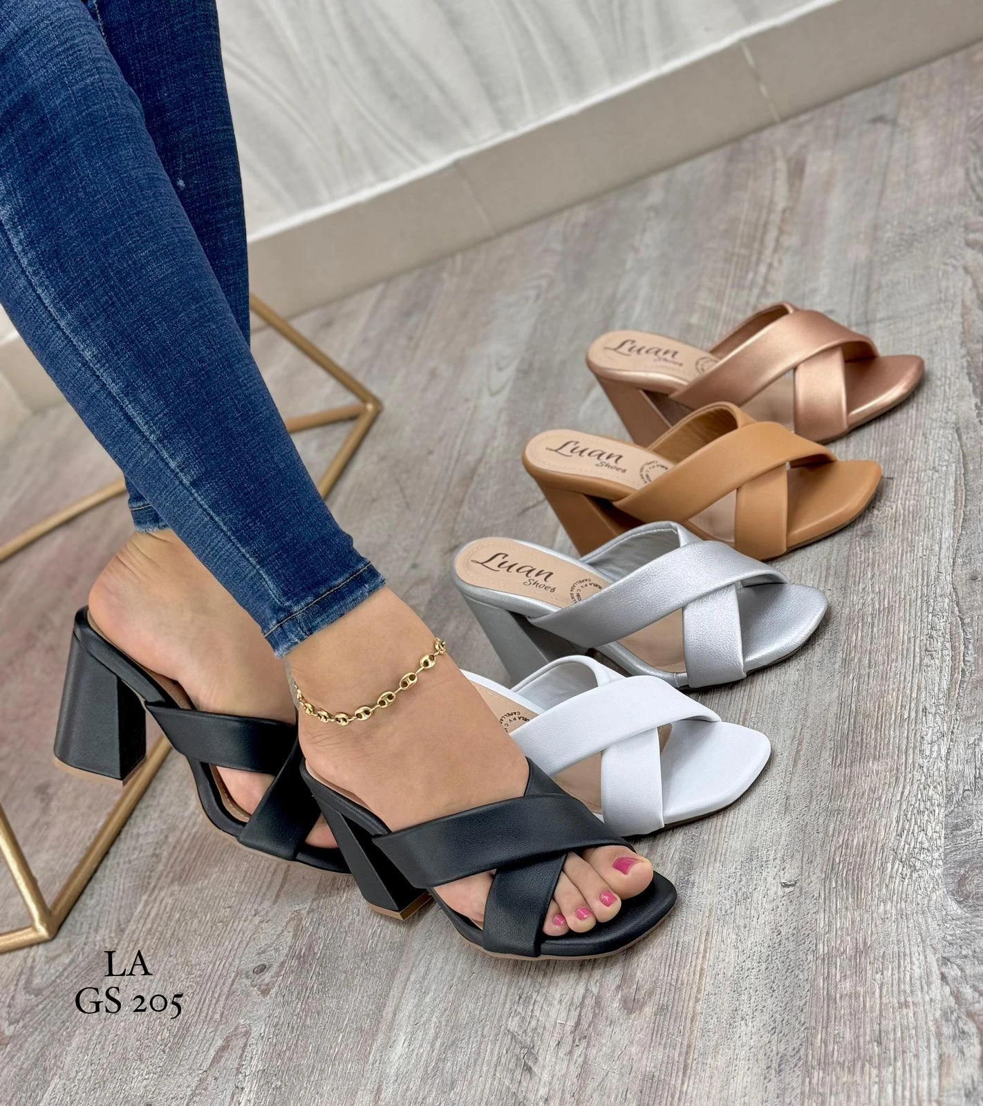 Sandalias de tacón cuadrado Nailin con tiras cruzadas, disponibles en tonos negro, blanco, plata, beige y bronce sobre fondo de madera.