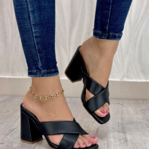 Sandalias de tacón cuadrado negras con diseño de tiras cruzadas en el empeine, lucidas con jeans y tobillera dorada sobre piso de madera clara.