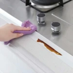 Mano limpia con paño morado borde blanco de estufa. Derrame café detenido por la cinta selladora.