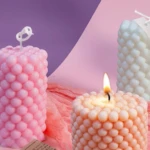 Velón redondo de silicona rosa con textura de burbujas, llama encendida y otros velones decorativos al fondo.