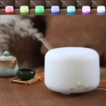 Humificador difusor aromaterapia blanco con vapor saliendo, taza té y tetera verde en mesa bambú.