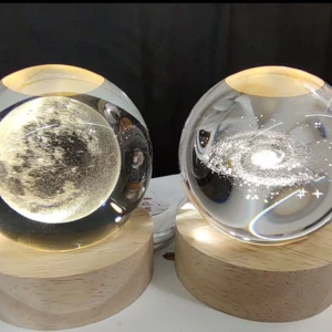 Dos esferas de cristal con grabados internos de la luna y una galaxia, sobre bases de madera clara con luz cálida LED para decoración de interiores.