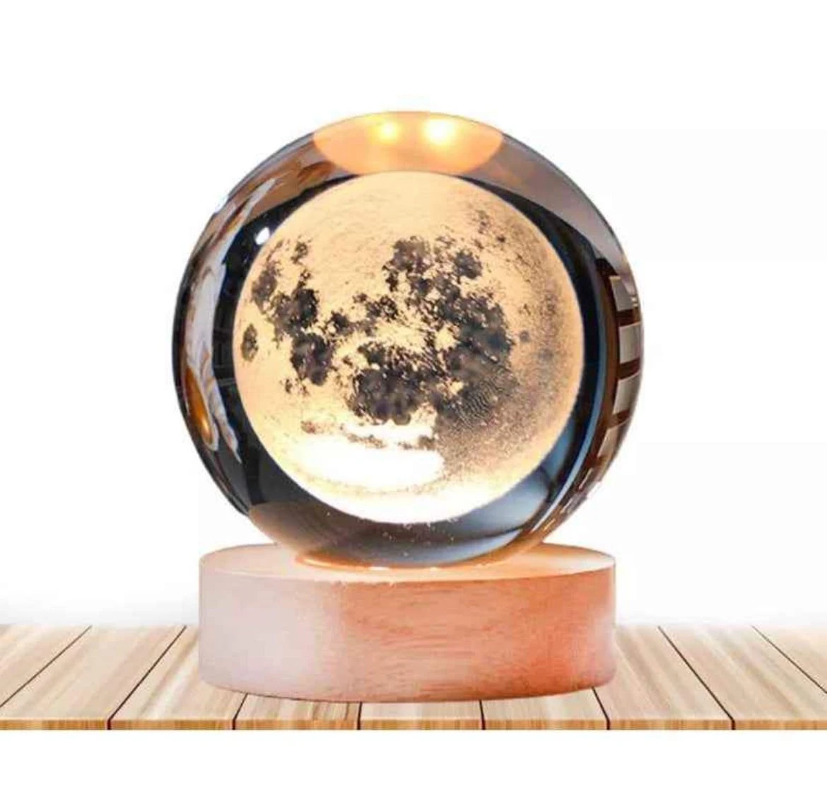 Lámpara esférica de cristal con grabado láser de la luna, sobre base cilíndrica de madera clara, iluminada con luz cálida sobre superficie de madera.