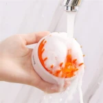 Cepillo de silicona para shampoo blanco con puntas naranjas, en uso bajo chorro de agua con espuma.