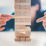 Torre de bloques de madera clara y jaspeada de Jenga con dedos extrayendo una pieza.