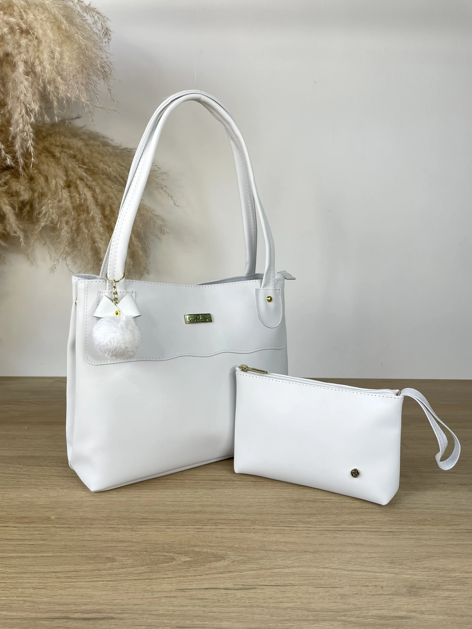Bolso tote blanco sintético Karla Chacon Gema, asa doble, con llavero pompón blanco y neceser a juego sobre mesa de madera.