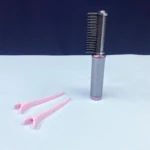 Cepillo alisador inalámbrico recargable color gris metalizado junto a dos pinzas de sección de cabello color rosa, sobre una superficie azul claro.