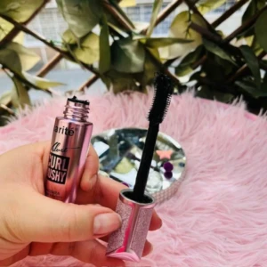 Pestañina Karité Curl Bushy rosa brillante, cepillo negro rizado, en mano sobre fondo rosa.