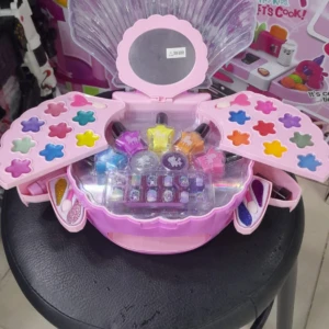 Set de maquillaje infantil en estuche rosa con forma de concha desplegable, incluye sombras, esmaltes y aplicadores organizados sobre un banco negro.