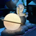 Lámpara de noche con astronauta sentado en luna y esfera luminosa simulando planeta.