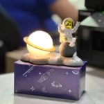 Lampara LED Astronauta sobre luna y nube, base púrpura con estrellas y cohete.