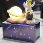 Lámpara de astronauta sobre luna con planeta y caja espacial, ideal decoración.