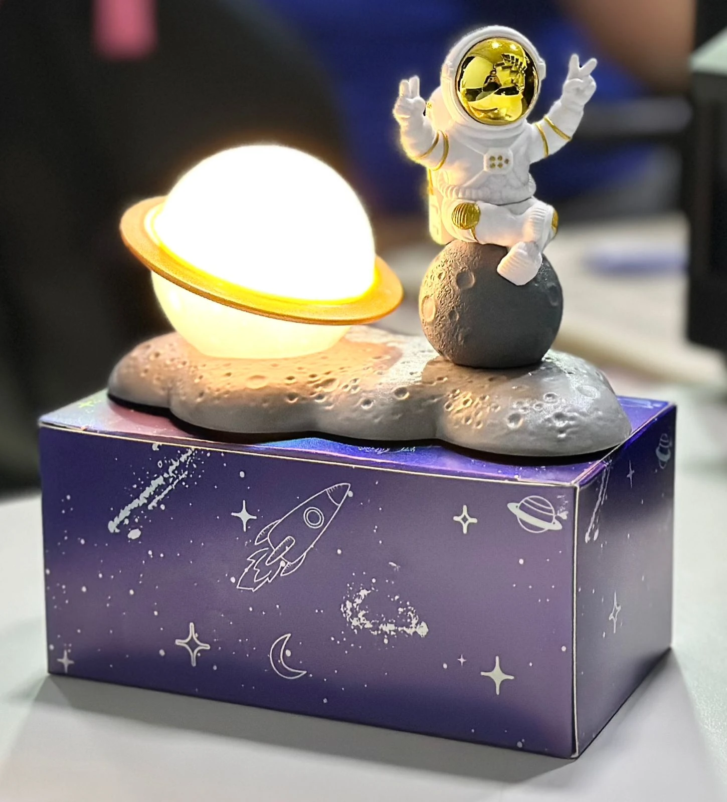 Lámpara de astronauta sobre luna con planeta y caja espacial, ideal decoración.