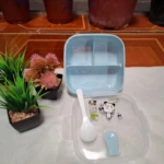 Lonchera cuadrada azul pequeña de plástico con dos divisiones y tapa transparente con dibujos de oso y cuchara.