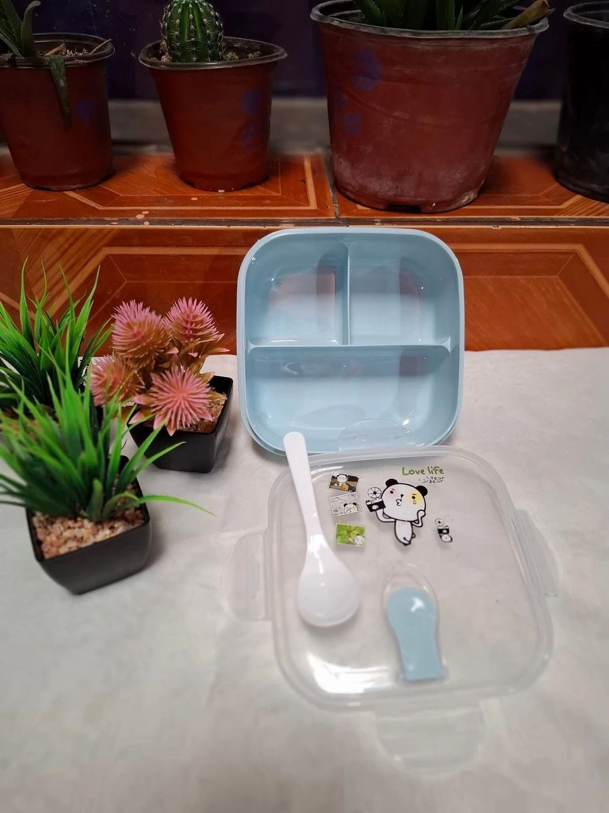 Lonchera cuadrada azul pequeña de plástico con dos divisiones y tapa transparente con dibujos de oso y cuchara.