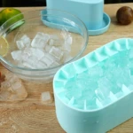 Molde de silicona celeste para cubos de hielo con cubos llenos y otros esparcidos en cuenco de cristal.