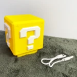 Lámpara en forma de bloque de interrogación Super Mario amarillo, con luces LED y cable USB, sobre superficie texturizada gris.