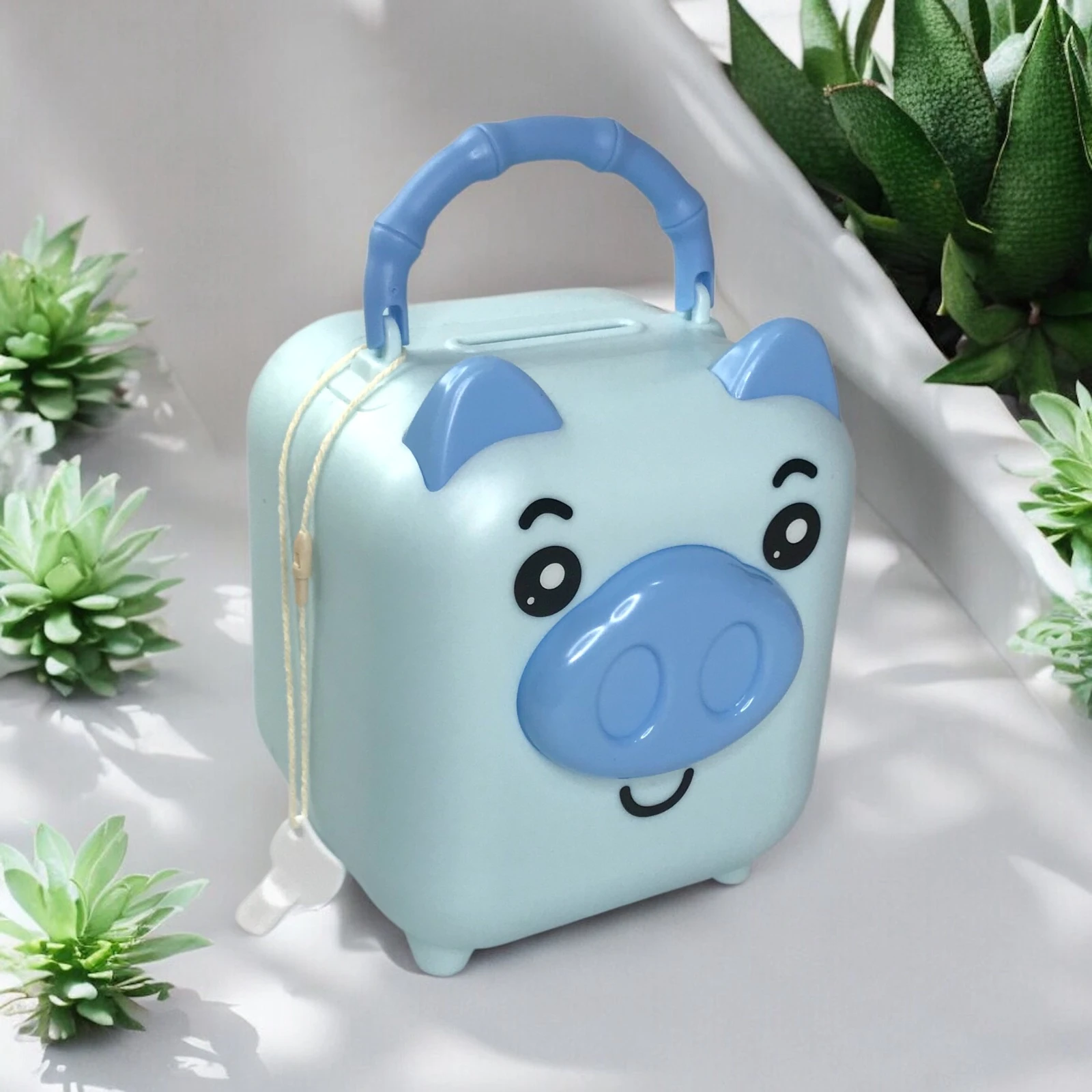 Alcancia azul con forma de cerdito, asa y llave, rodeada de suculentas.