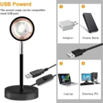 Lámpara efecto atardecer con base circular y cable USB, junto a íconos que muestran su compatibilidad con adaptadores, bancos de energía, laptops y PC.