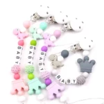 Cadenas para chupo con dinosaurios, coronas y letras "BABY" en tonos pastel y gris.