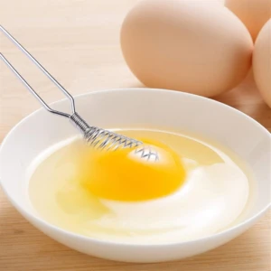 Batidor manual de resorte de acero inoxidable batiendo un huevo en un tazón blanco.