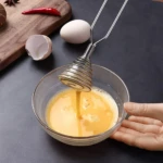 Batidor manual de resorte metálico batiendo huevo en un bol de vidrio.