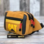 Canguro Caterpillar color mostaza con logo amarillo, dos cierres con cordón rojo, sobre caja de presentación.