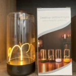 Lámpara táctil imantada de luz cálida con base negra y domo de vidrio transparente junto a su caja de empaque con diseño de modelos de colores variados.