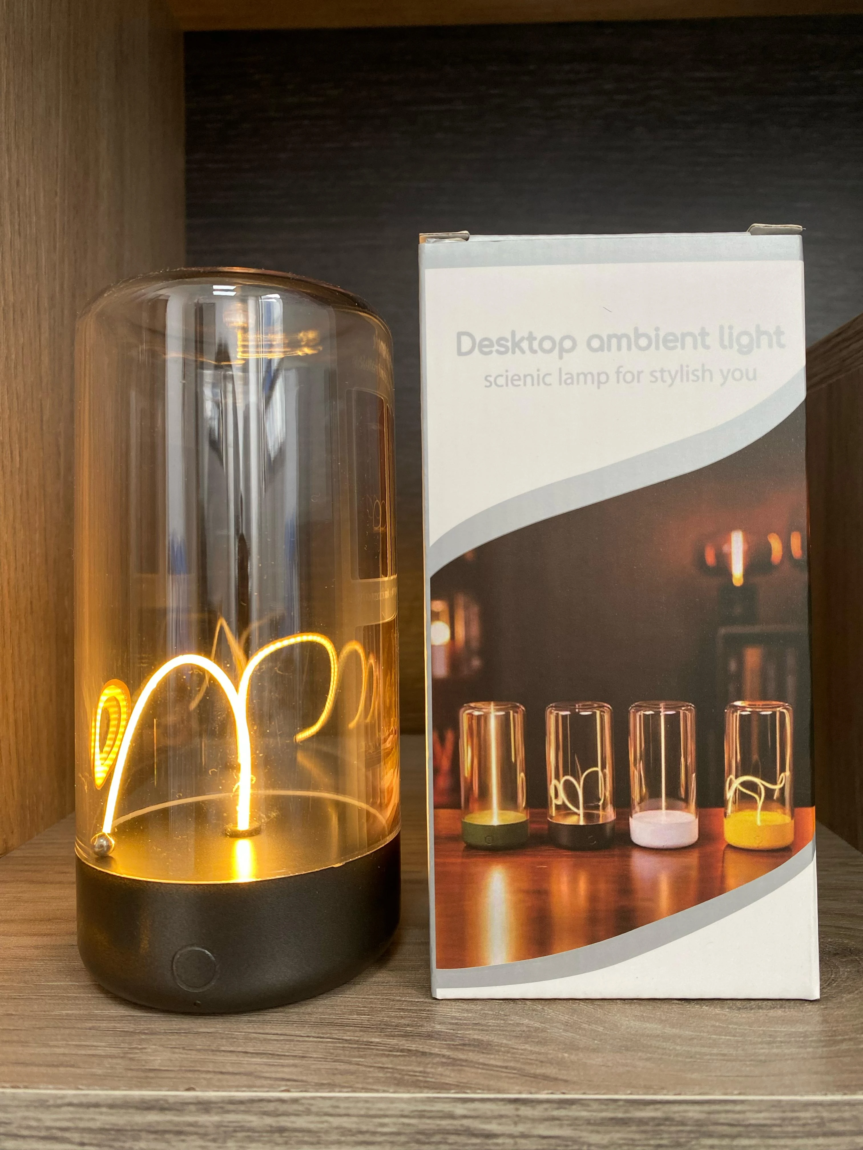 Lámpara táctil imantada de luz cálida con base negra y domo de vidrio transparente junto a su caja de empaque con diseño de modelos de colores variados.