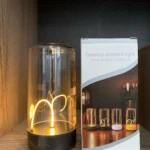 Lámpara táctil imantada de sobremesa con cuerpo de vidrio cilíndrico y filamento luminoso curvo, junto a su caja de empaque con diseño decorativo.
